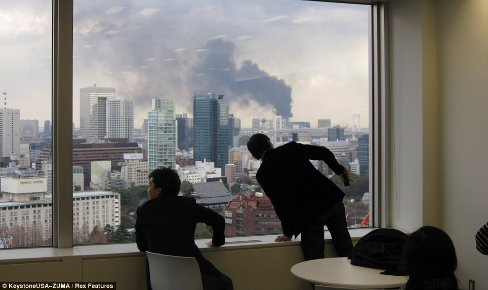 Concerned: Two office workers in Tokyo brace themselves for a potential disaster as the earthquake rumbled buildings and smoke can be seen rising from skyscrapers outside